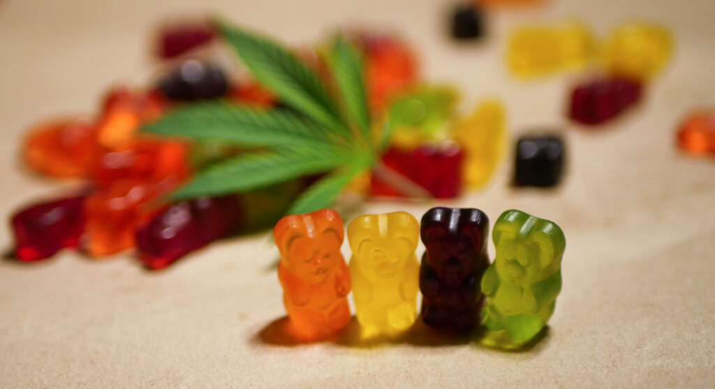 Close-up of colorful gummy bears with a blurred green leafy plant in the background, warm neutral tones, relaxed atmosphere